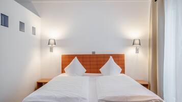 Cama doble con sábanas blancas y dos almohadas, flanqueada por mesitas de noche con lámparas, en una habitación con pared blanca lisa y un cabecero naranja estampado.
