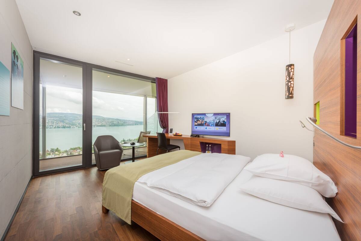 Ein Hotelzimmer mit Bett, Schreibtisch, Stuhl, Fernseher und einem großen Fenster mit Blick ins Freie.