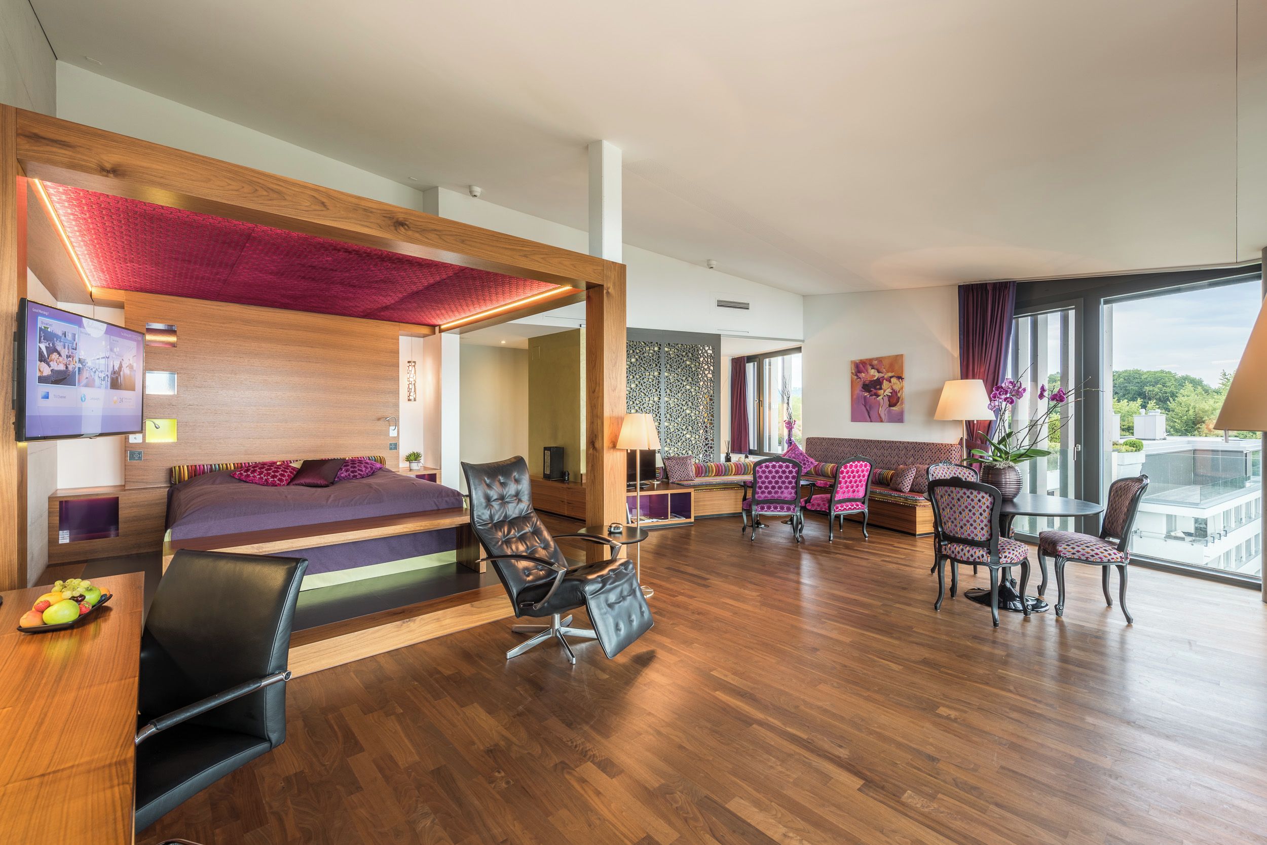 A modern hotel room with a wooden floor, a bed with a red canopy, a seating area with patterned chairs, and a large window offering a view.
