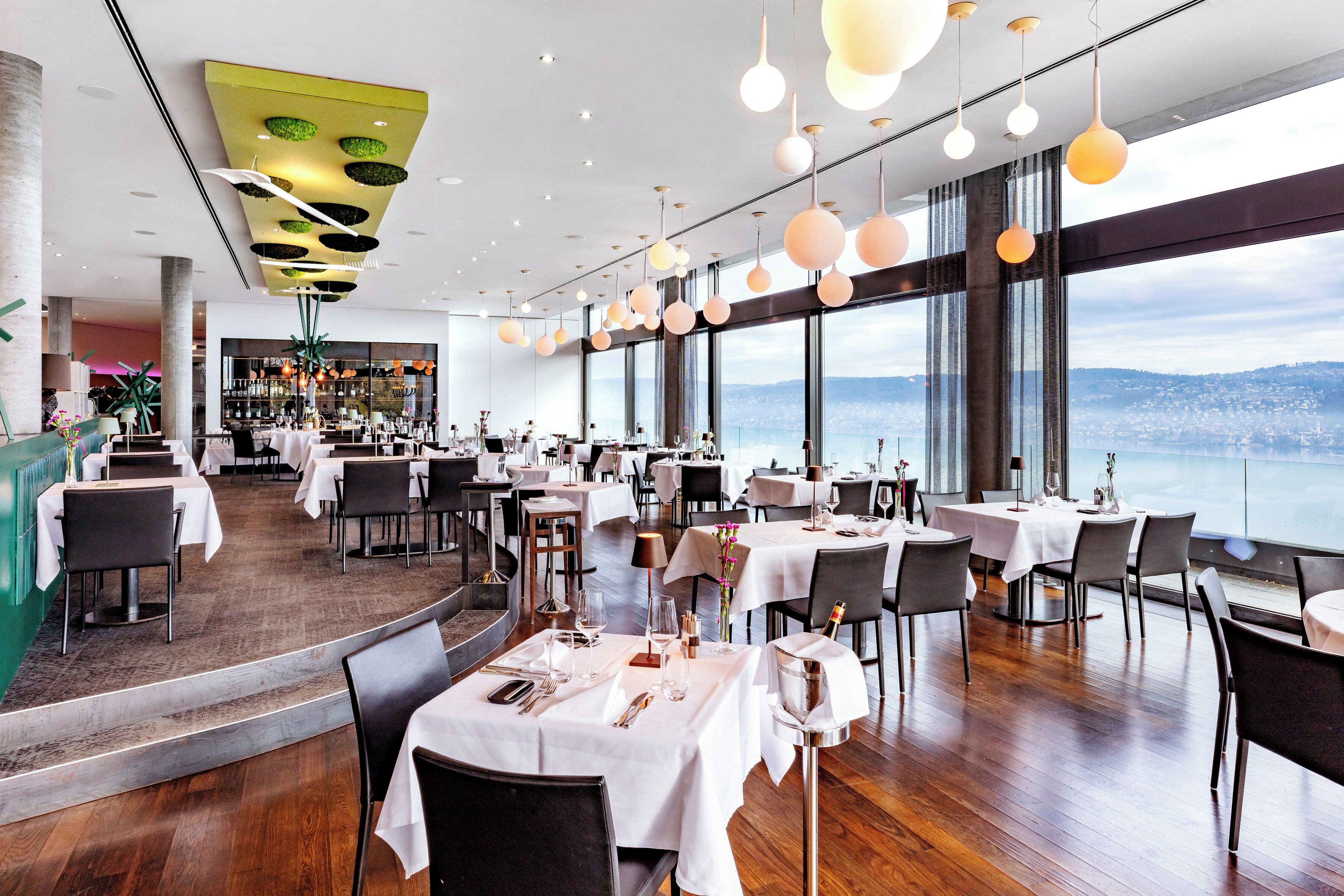 A restaurant with tables set for dining, large windows offering a city view, and hanging spherical light fixtures.