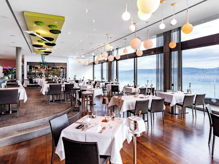 A restaurant with tables set for dining, large windows offering a city view, and hanging spherical light fixtures.