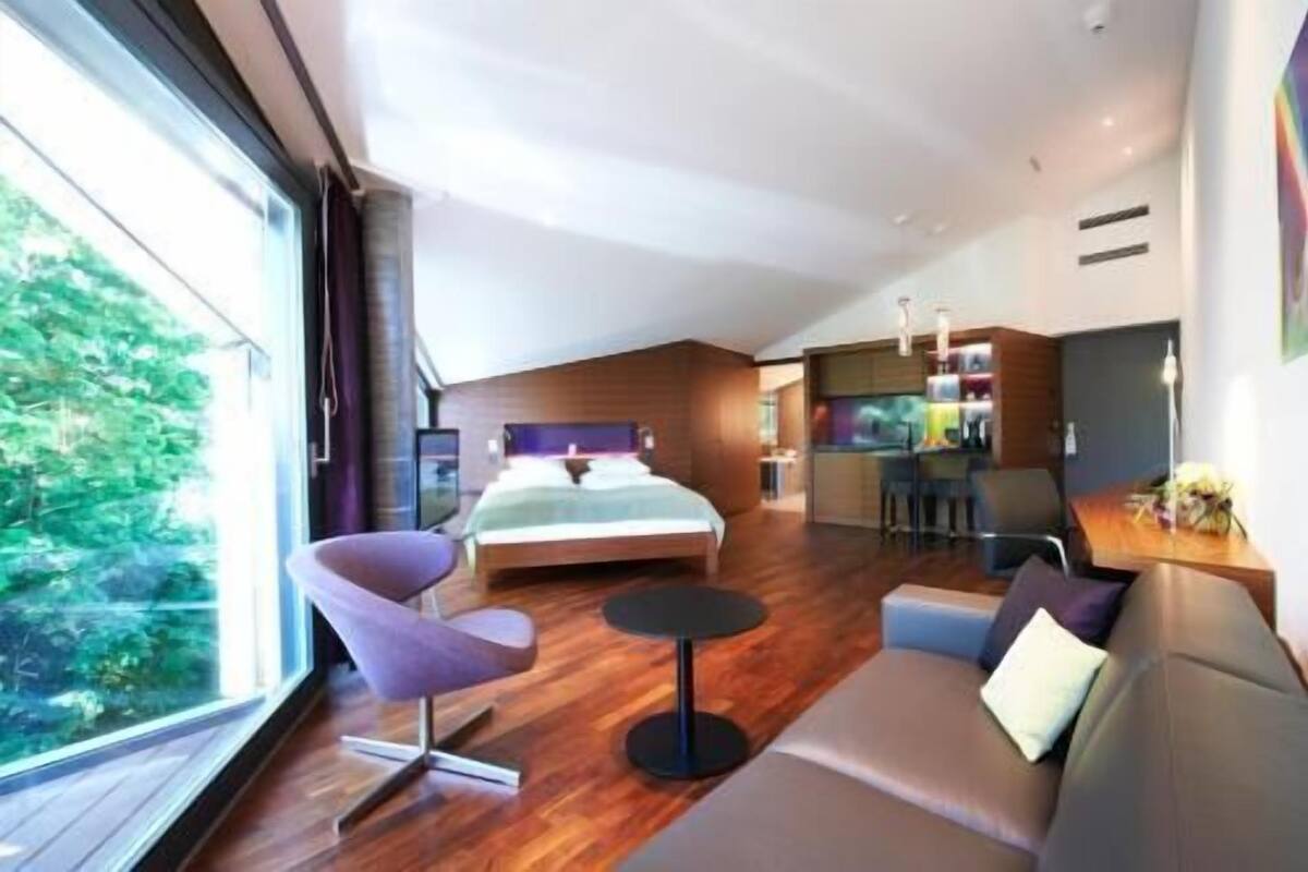 Ein modernes Hotelzimmer mit einem Bett, einer Sitzecke inklusive Sofa und Sesseln, einem kleinen Tisch und Blick auf Grünflächen.