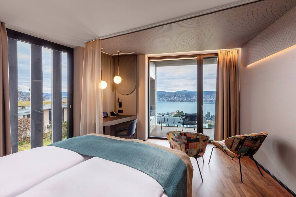 Ein modernes Hotelzimmer mit einem großen Bett, einem Schreibtisch, einem Sessel und einem Balkon mit Blick auf die Stadt und das Wasser.