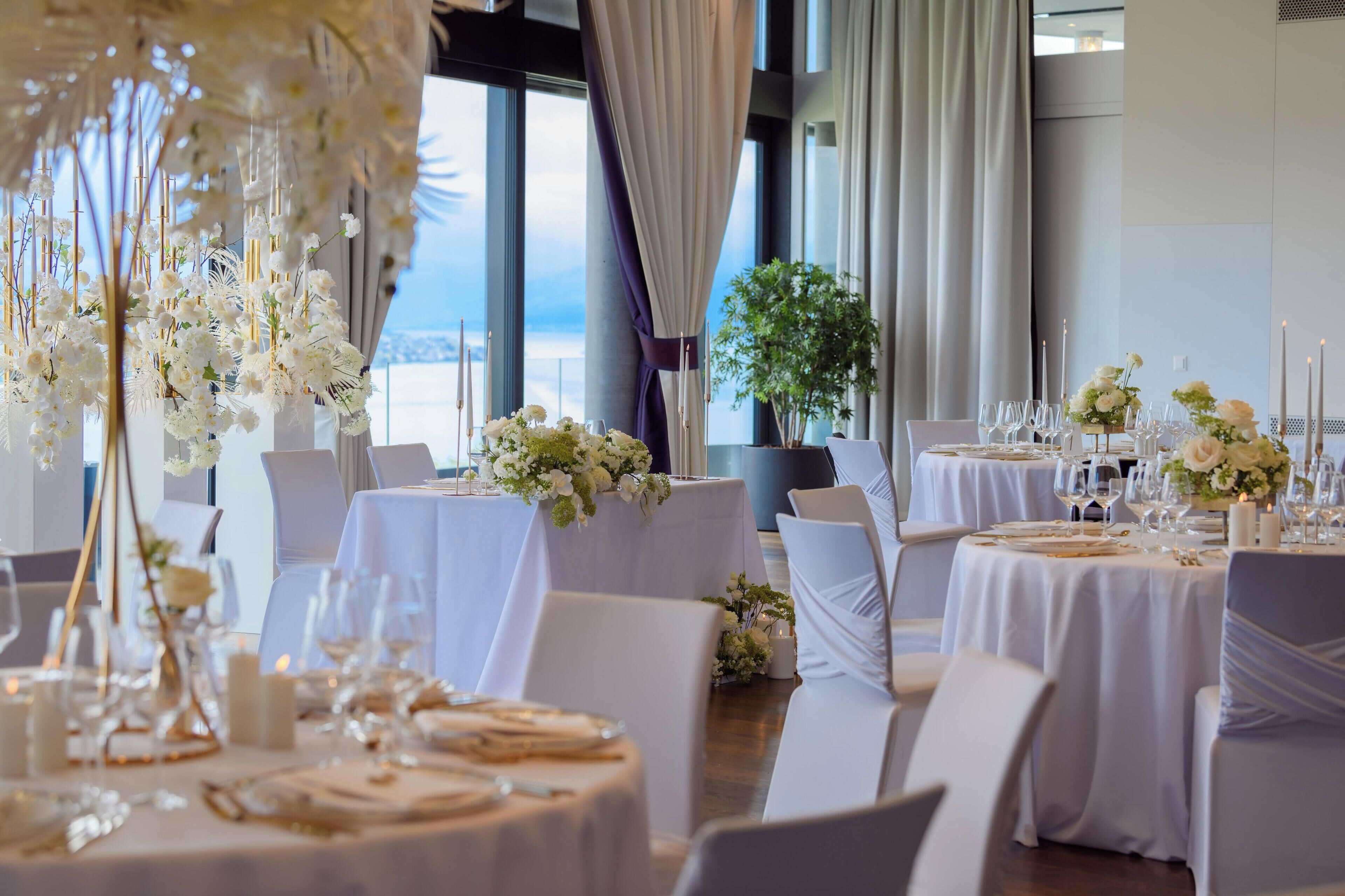Une salle de banquet avec des tables rondes dressées pour le repas, des tentures blanches et des compositions florales.