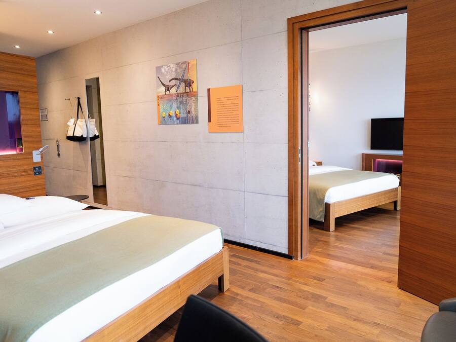 A hotel room with two beds, a wooden floor, a wall-mounted TV, and a door leading to another room.
