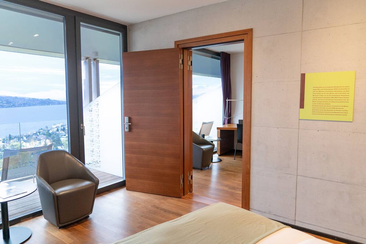 Ein modernes Hotelzimmer mit einem großen Fenster, das einen Blick auf die Stadt bietet, einer Holztür, einem braunen Ledersessel und einem kleinen Tisch.