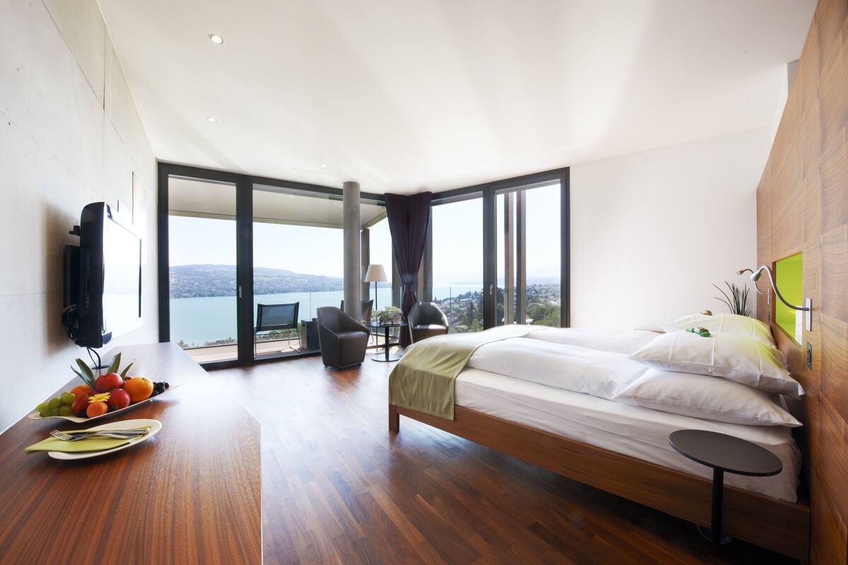Ein modernes Hotelzimmer mit einem großen Bett, einem Esstisch mit Früchten, einem Fernseher und Blick ins Freie.