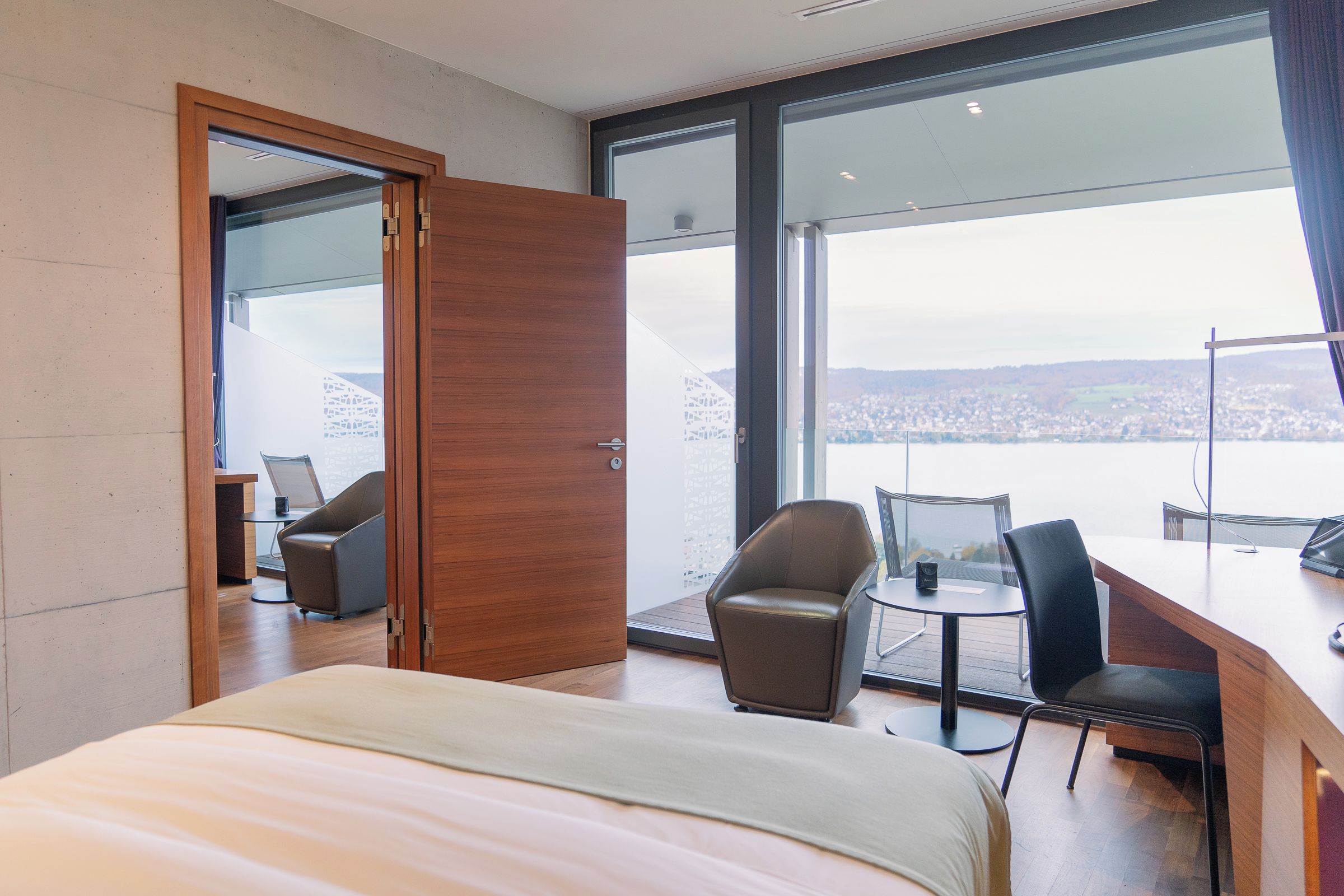 Een hotelkamer met een groot bed, twee stoelen, een klein tafeltje en een stadsgezicht door grote ramen.