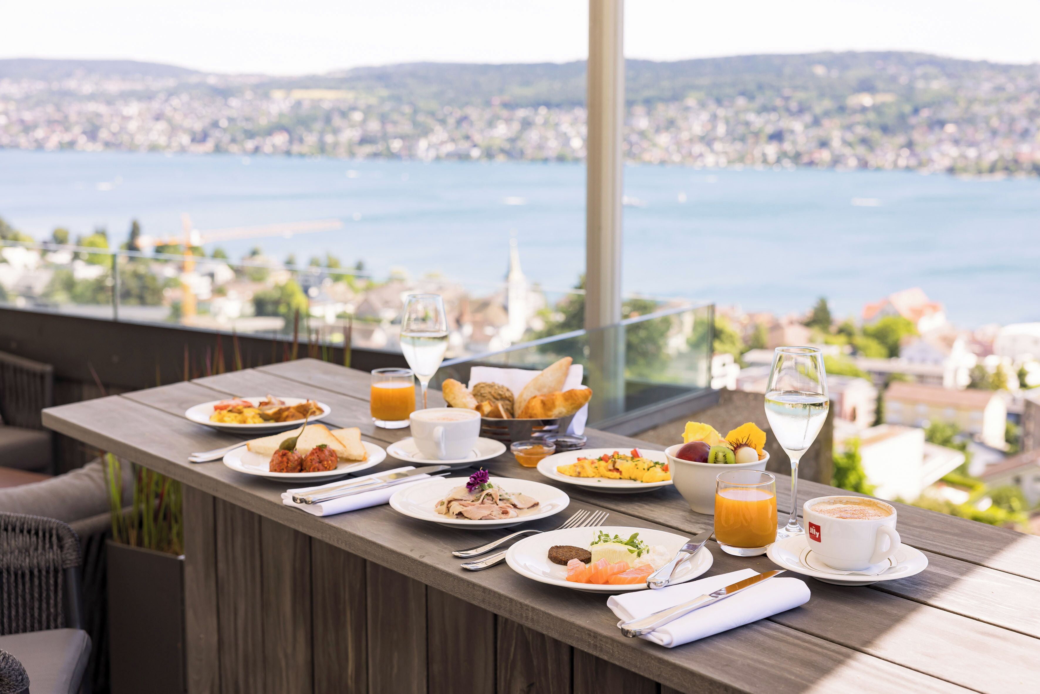 A breakfast table with various dishes, a glass of orange juice, and a view of a city and water.