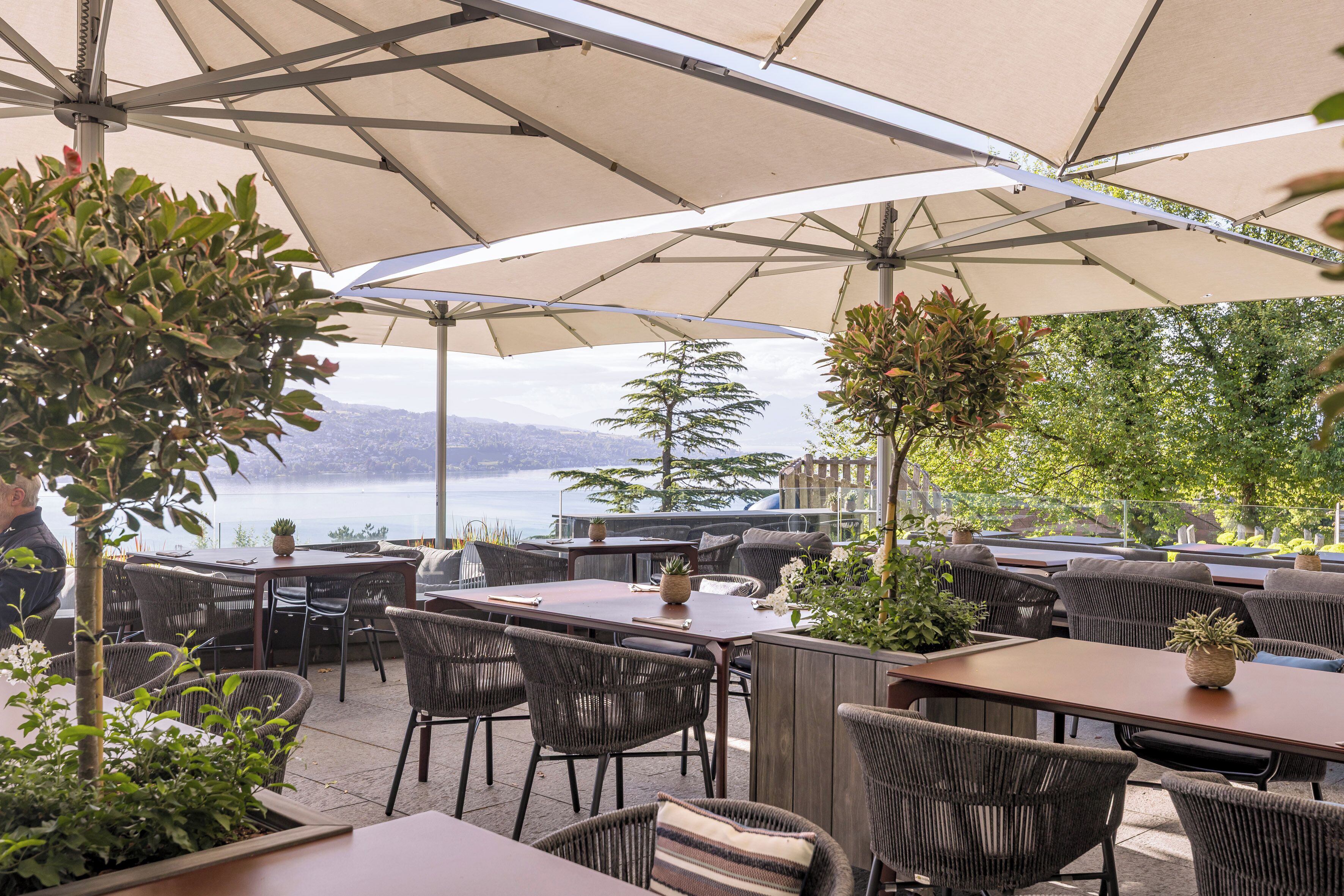 An outdoor seating area with tables and chairs, a large umbrella, and a view of the lake.