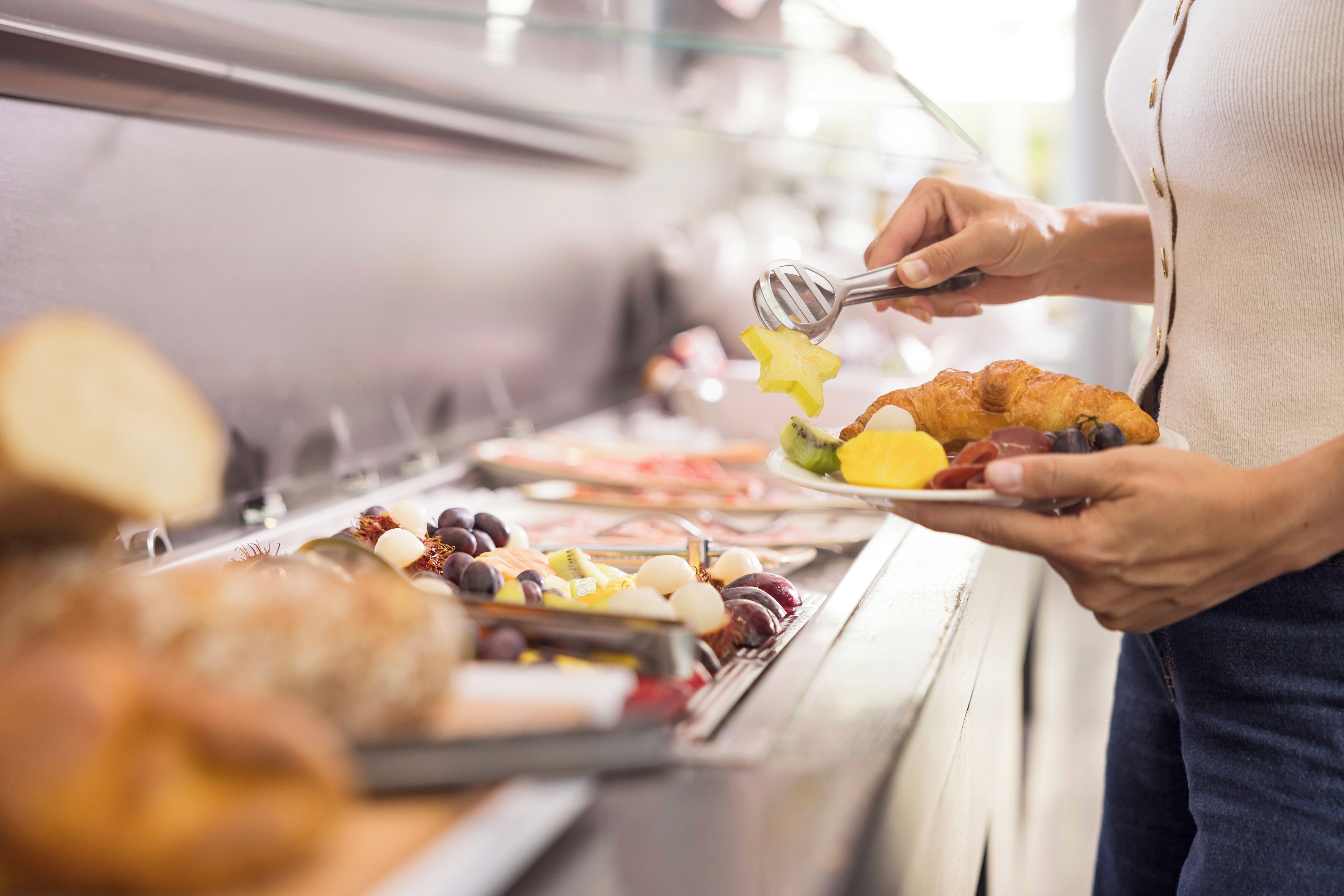 Une personne tenant une assiette remplie de différents aliments, notamment un croissant et des fruits, devant un buffet.