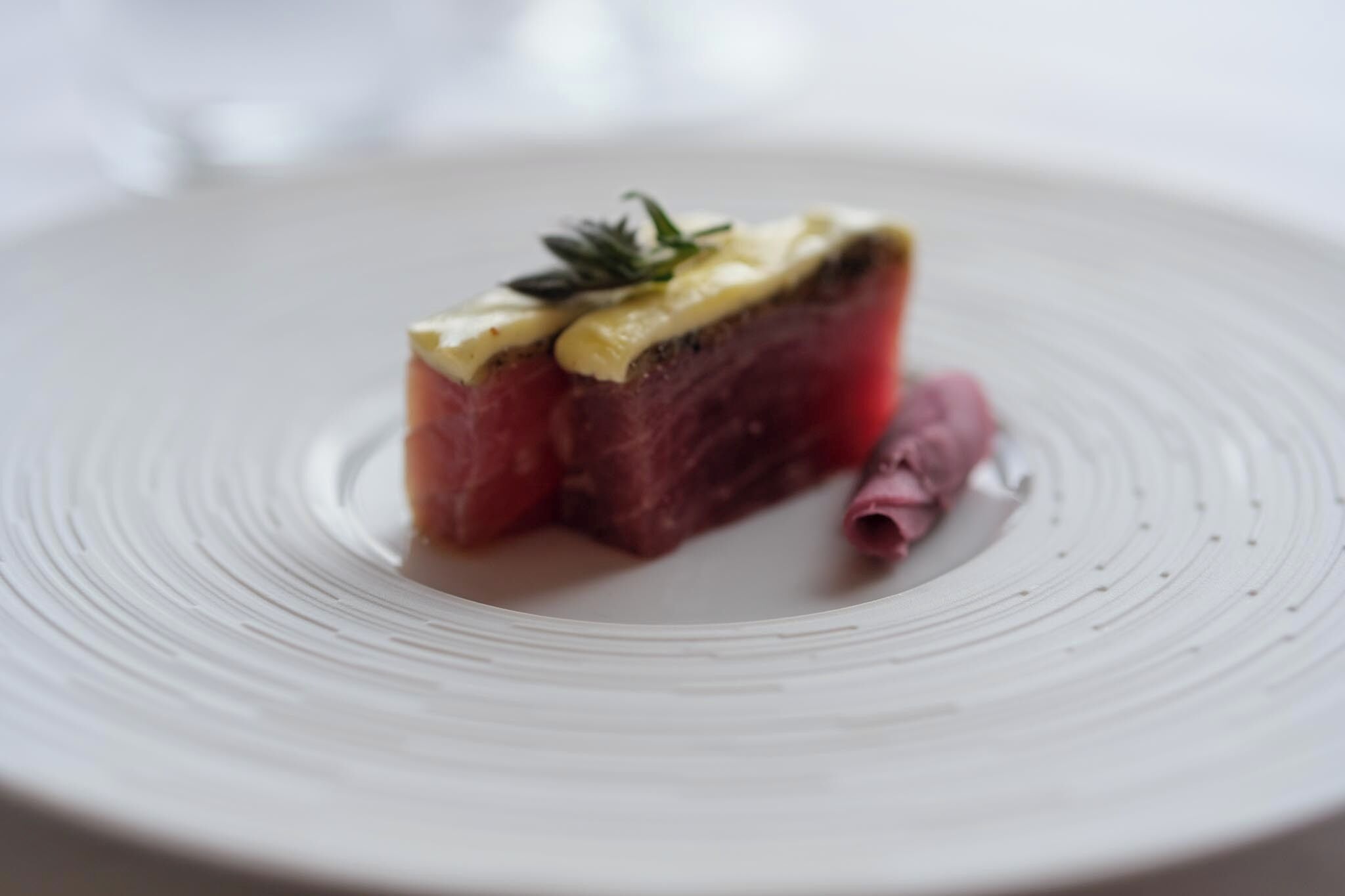 Une assiette contenant une tranche de viande recouverte d’une sauce crémeuse et agrémentée d’une branche d’herbe aromatique.