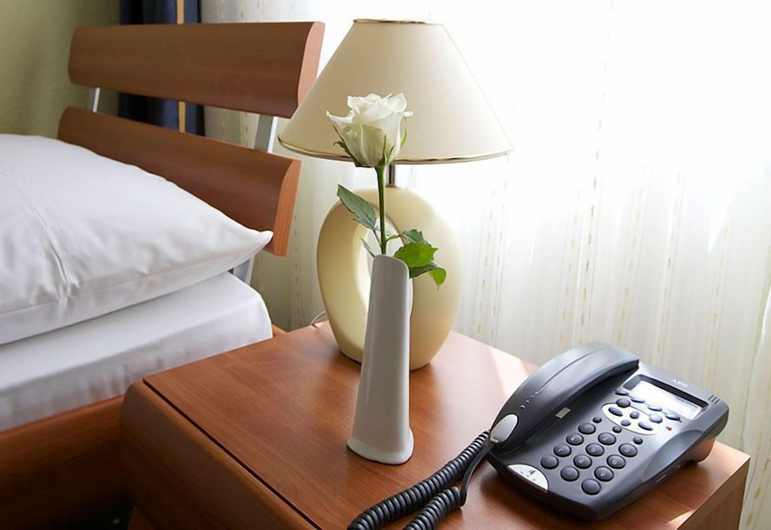 Una habitación de hotel con una cama, una mesita de noche con lámpara, un jarrón con una flor y un teléfono.