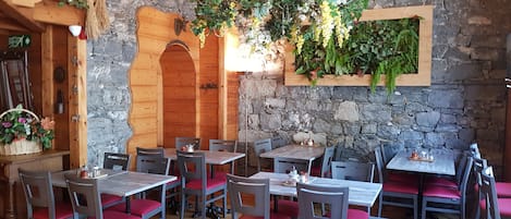 A cozy restaurant with wooden tables and chairs, stone walls, and hanging plants.