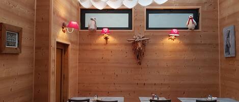 A wooden interior with a long table and chairs, a skull mounted on the wall, and two wall-mounted lamps.