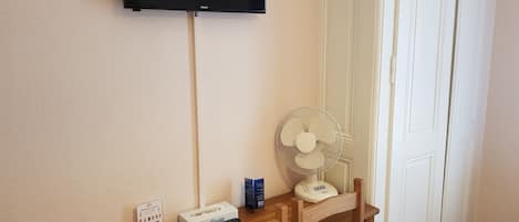 A room with a flat-screen TV mounted on the wall, a standing fan, a wooden table with a remote control, a tissue box, and a sign indicating a minibar.