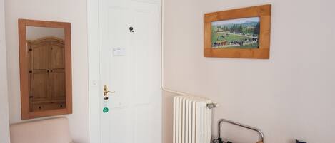 A room with a white door, a radiator, a wooden chair, a small trash bin, and a framed picture on the wall.