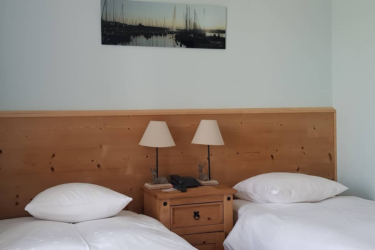 A hotel room with a wooden headboard, two beds with white bedding, two bedside lamps, a nightstand with a phone and a small statue, and a picture of a marina on the wall.