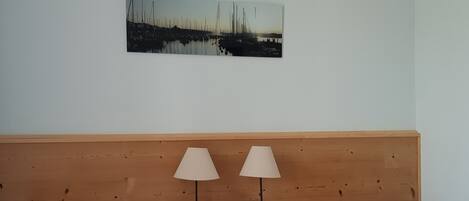 Une chambre d’hôtel avec une tête de lit en bois, deux lits avec une literie blanche, deux lampes de chevet, une table de chevet avec un téléphone et une petite statue, et un tableau représentant un port de plaisance accroché au mur.