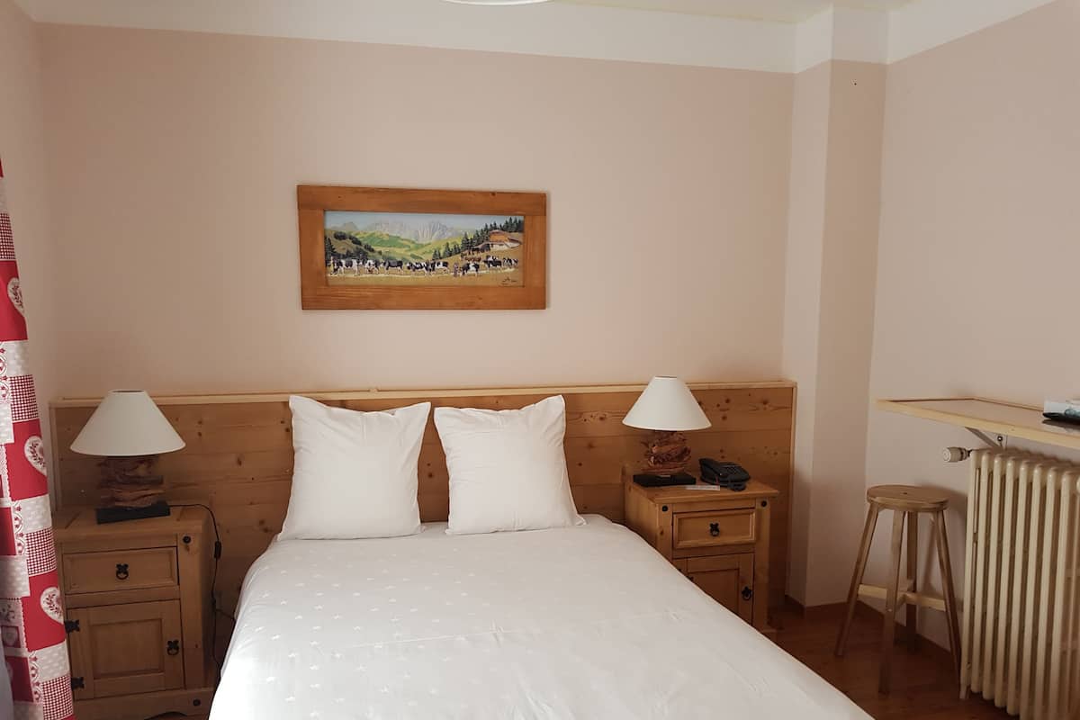 A bedroom with a bed, two bedside tables with lamps, a wooden headboard, a painting on the wall, a radiator, and a wooden stool.