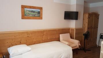 Un lit simple avec un matelas et un oreiller blancs, une tête de lit en bois, un téléviseur fixé au mur, une armoire en bois et un ventilateur sur pied.
