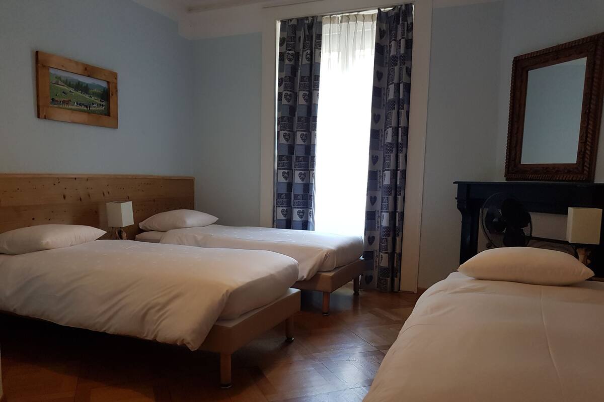 A hotel room with two beds, a wooden headboard, a bedside table with a lamp, a large mirror, and a window with patterned curtains.