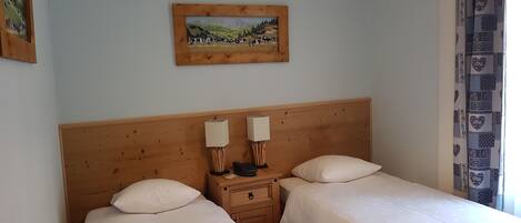 A room with two single beds, wooden headboards, bedside tables with lamps, a small drawer, and a framed painting on the wall.