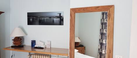 Una habitación con un escritorio de madera, dos taburetes, un radiador, una cama con cortinas estampadas, un cuadro en la pared y un espejo.