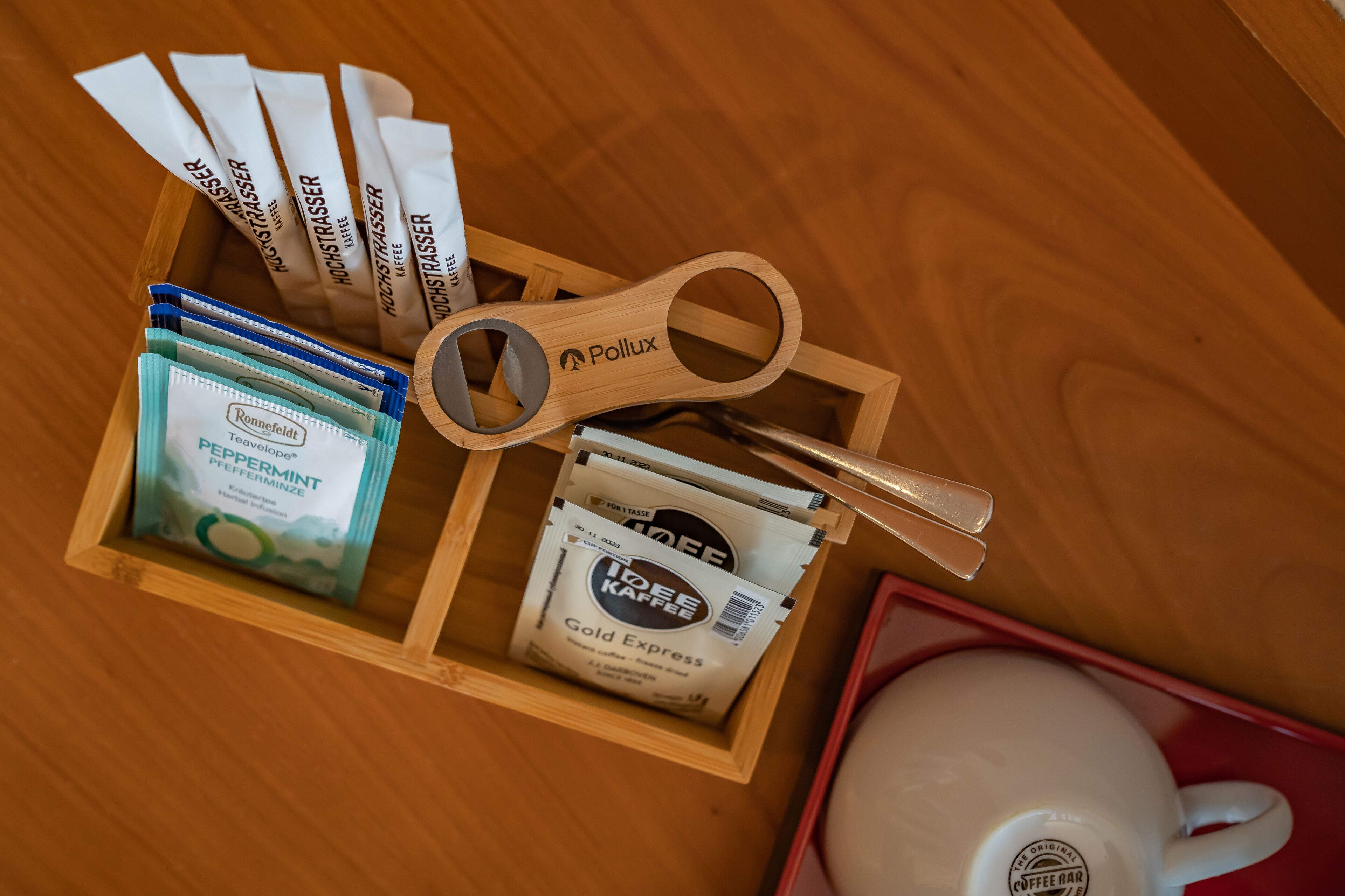 Una bandeja de madera con sobres de café, una cuchara de madera y una taza blanca sobre una superficie de madera.