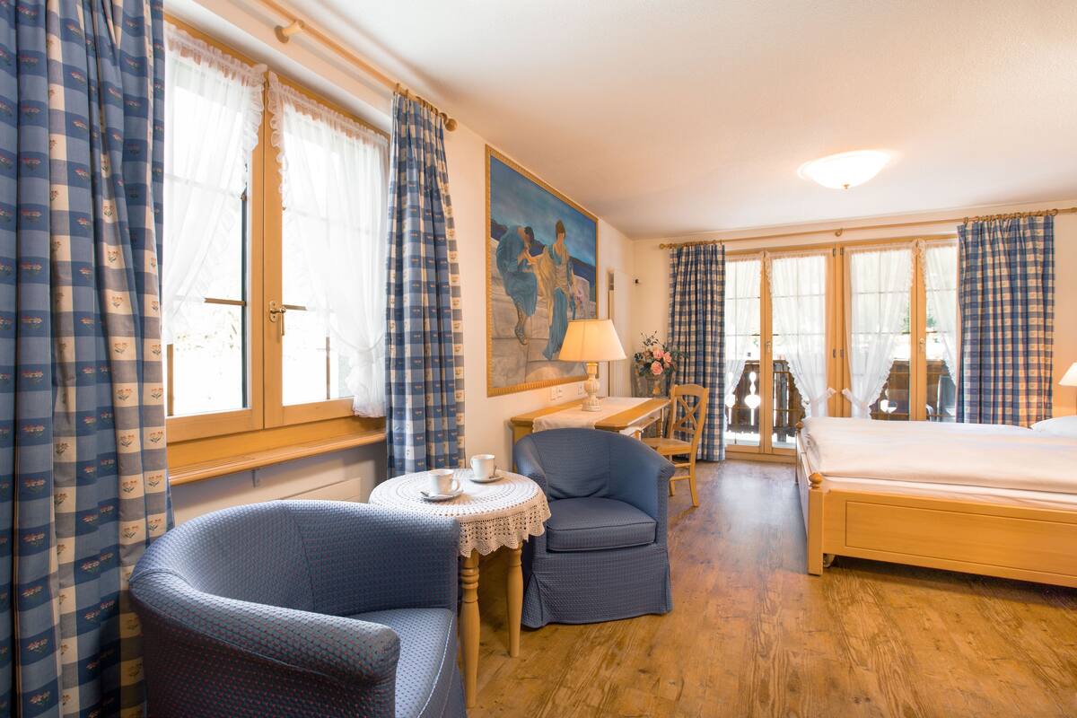 Ein Hotelzimmer mit Holzboden, einem Bett, einem Schreibtisch mit Lampe, zwei Stühlen, einem kleinen Tisch mit Tasse und Untertasse sowie Vorhängen mit Karomuster.