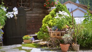 A rustic wooden house with a garden in front, including a wheelbarrow with plants and a small tree. - Hotel Romantica (Zermatt)