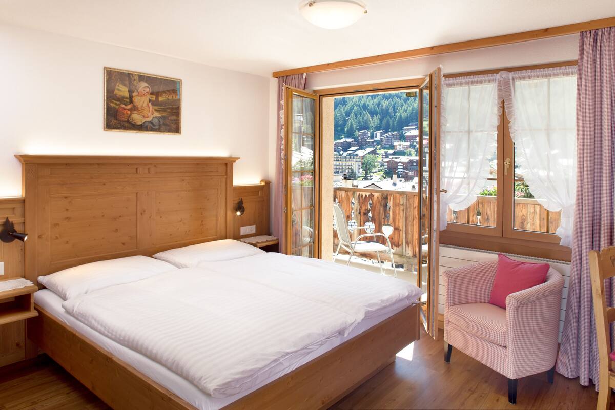 Ein Schlafzimmer mit einem hölzernen Bett, einem Sessel und einem Balkon mit Blick auf ein Dorf.