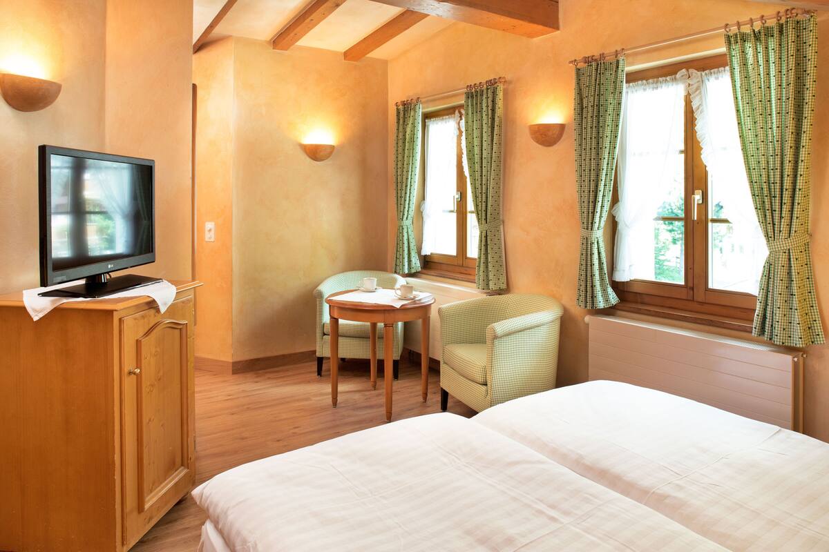 Ein Zimmer mit einem Bett, einem Fernseher auf einer Holzkommode, einem kleinen Tisch mit einem Stuhl und einem Fenster mit Vorhängen.