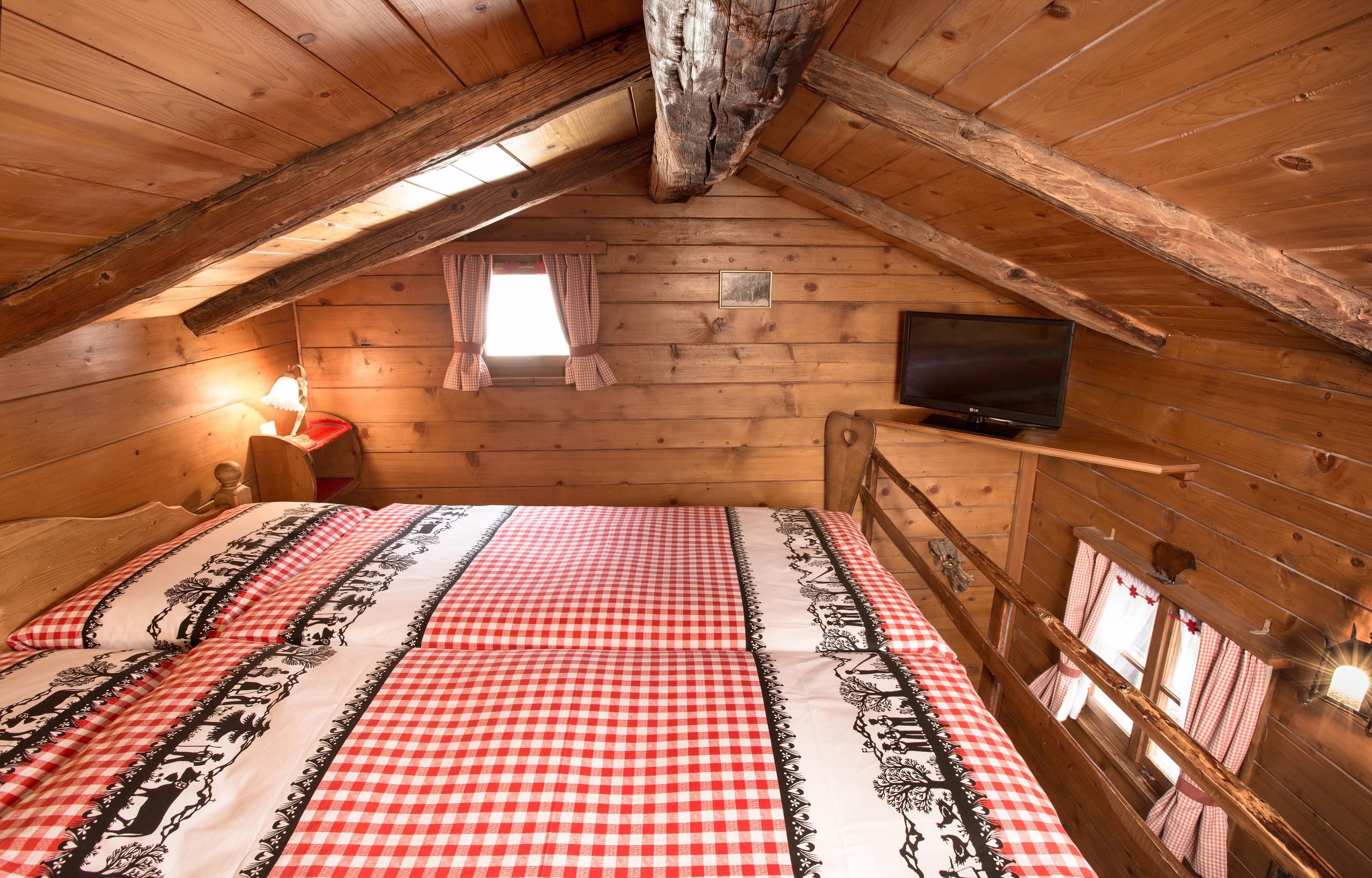 Un dormitorio acogedor en el ático, con colcha a cuadros, televisión y una ventana pequeña con cortinas.