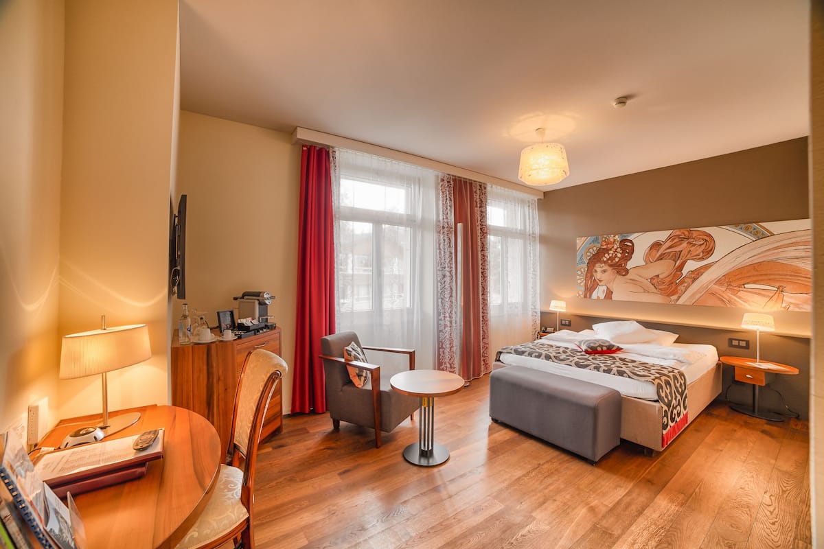 A hotel room with a bed, a desk with a lamp, a chair, a small table, and a painting on the wall.