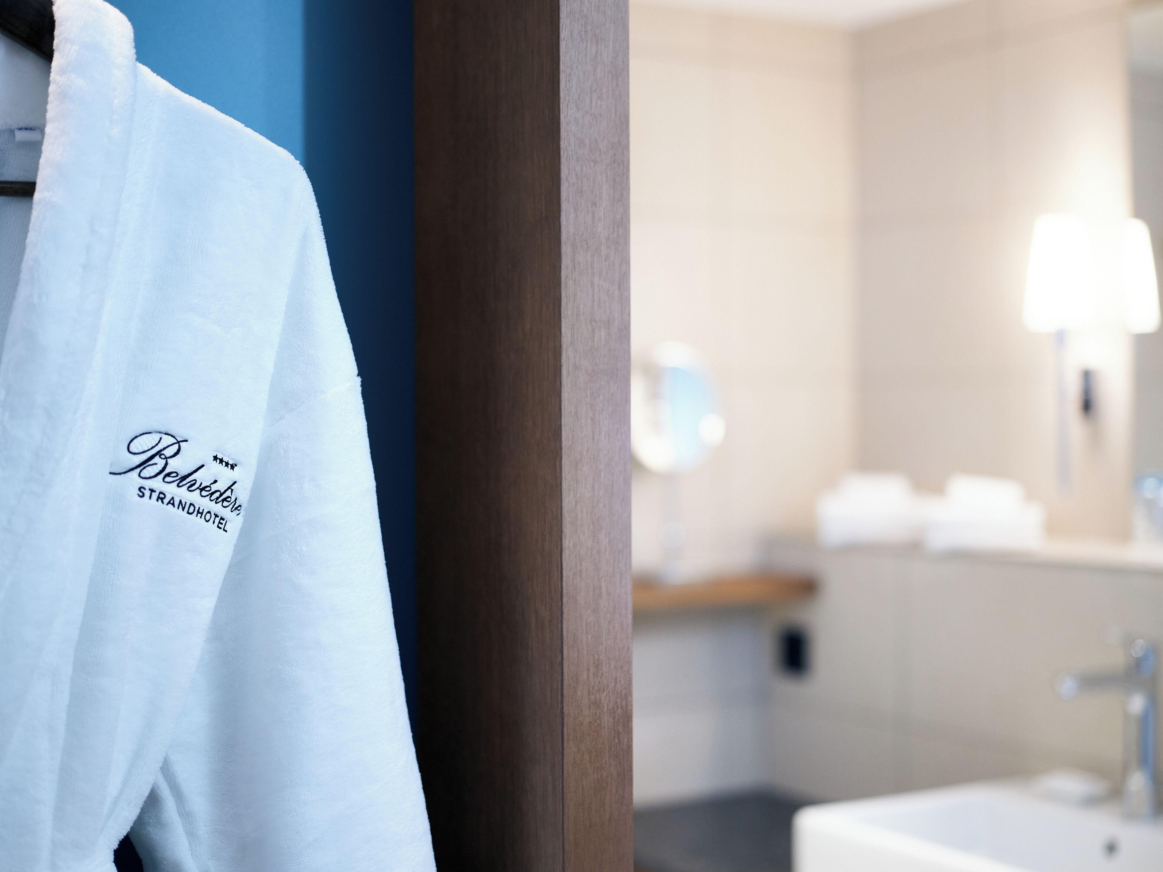 A hotel room with a white robe hanging on a wooden door, a bathroom with a sink and mirror, and a wooden shelf above the mirror.