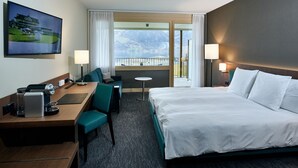 A hotel room with a bed, a desk with a chair, a small table, a TV, and a view of mountains and a lake. - Belvédère Strandhotel (Spiez)