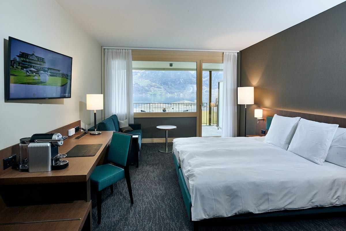 A hotel room with a bed, a desk with a chair, a small table, a TV, and a view of mountains and a lake.