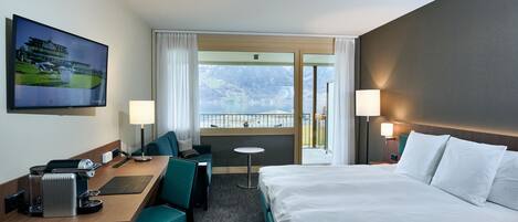 Una habitación de hotel con una cama, un escritorio con silla, una mesita, un televisor y vistas a montañas y un lago.