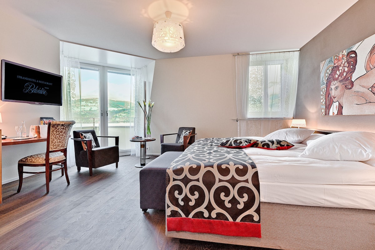 A hotel room with a bed, a desk with a TV, a chair, a table with a vase, and large windows.