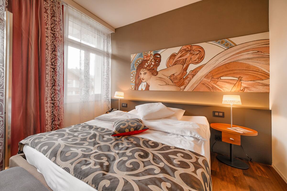 A hotel room with a bed, a nightstand, a lamp, a large painting on the wall, and a window with curtains.