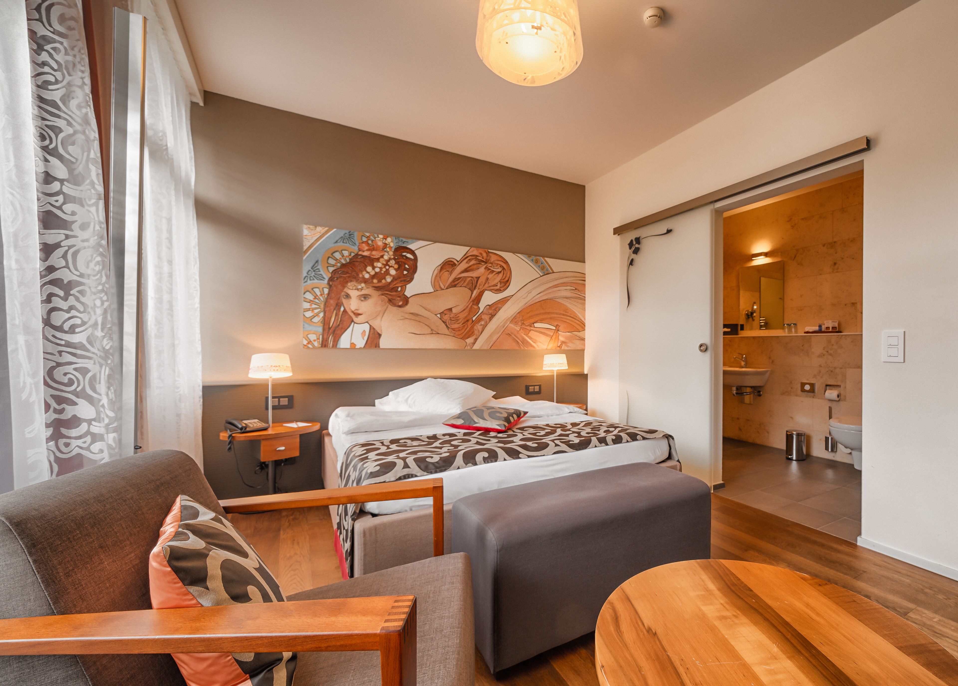 A modern hotel room with a large bed, a sitting area with a wooden bench, a painting on the wall, and a bathroom visible through an open door.