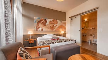 A modern hotel room with a large bed, a sitting area with a wooden bench, a painting on the wall, and a bathroom visible through an open door.