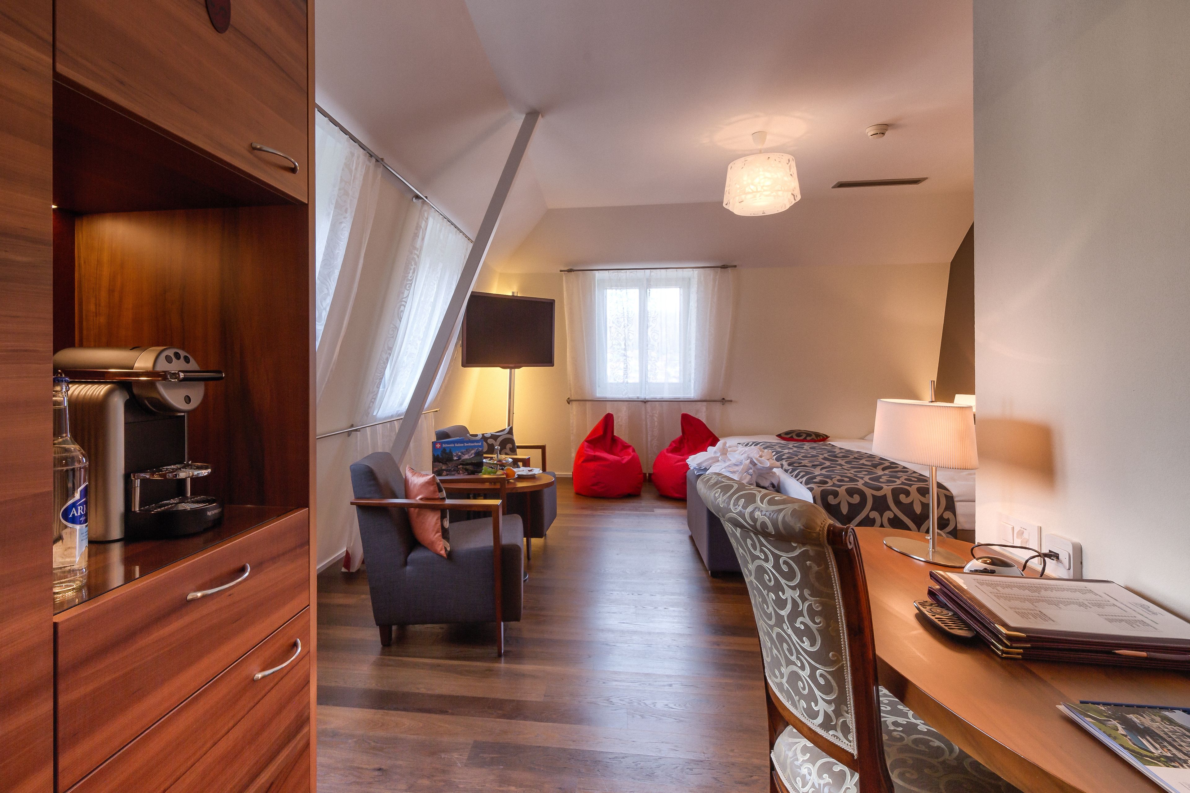Habitación de hotel compacta con armario de madera, un escritorio con lámpara, un sofá y una cama con cabecera estampada.
