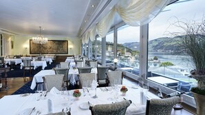 A restaurant with tables set for dining, large windows offering a view of a body of water and hills, and a chandelier hanging from the ceiling. - Belvédère Strandhotel (Spiez)