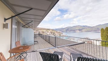 A balcony with a wooden table and chairs, overlooking a lake and distant hills.