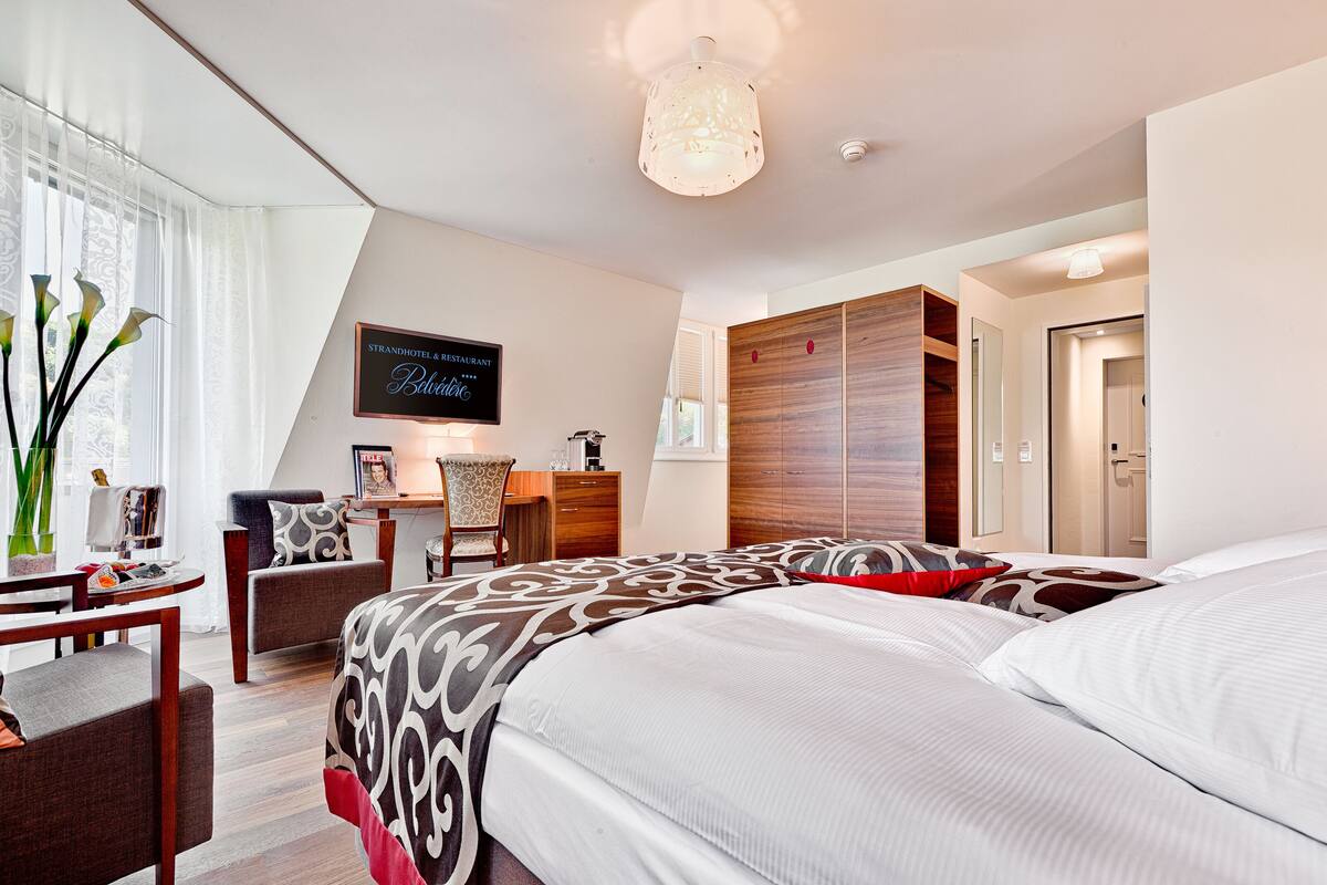 A modern hotel room with a large bed, a desk, a chair, and a wardrobe.