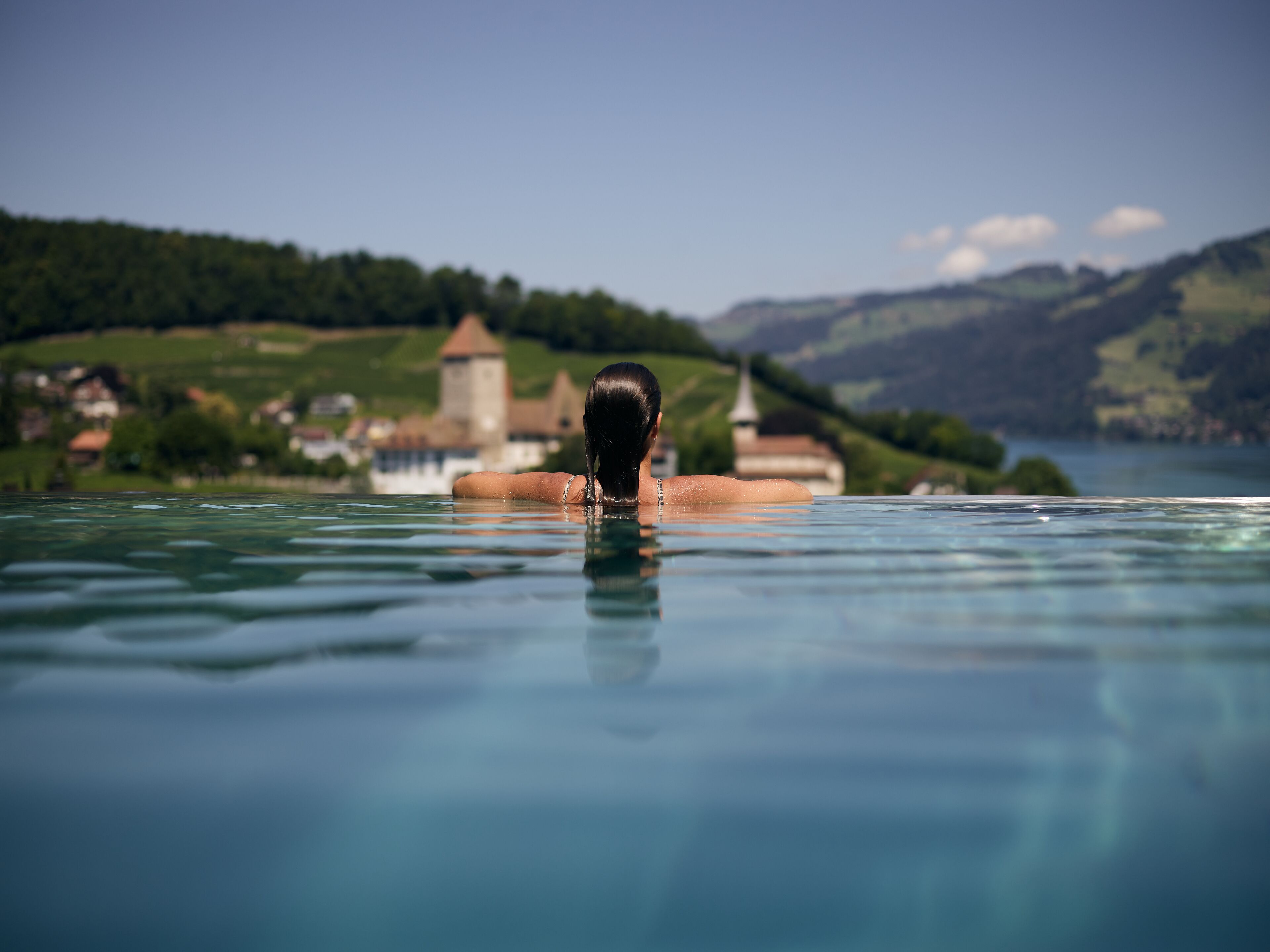 A person in a swimming pool with a view of a lake, mountains, and buildings in the background.