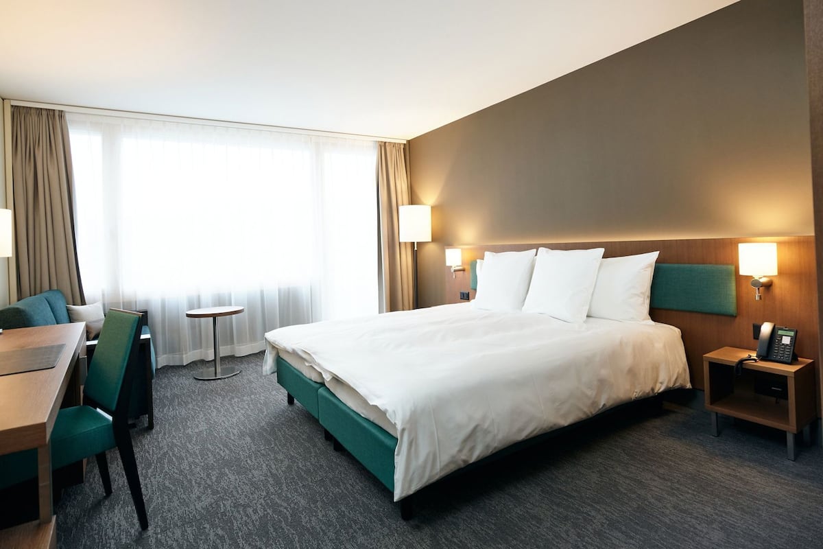 A hotel room with a bed, a desk, a chair, a small table, and a lamp.