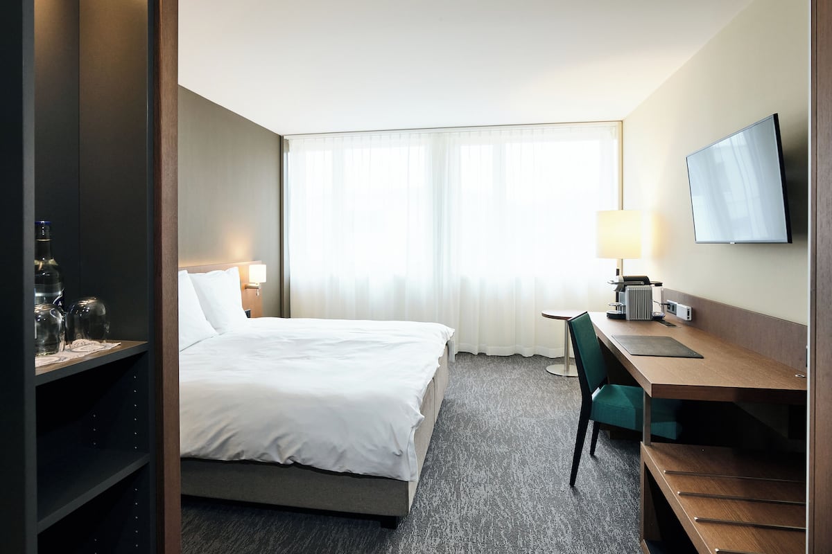 A hotel room with a bed, a desk, a chair, a TV, and a window with curtains.